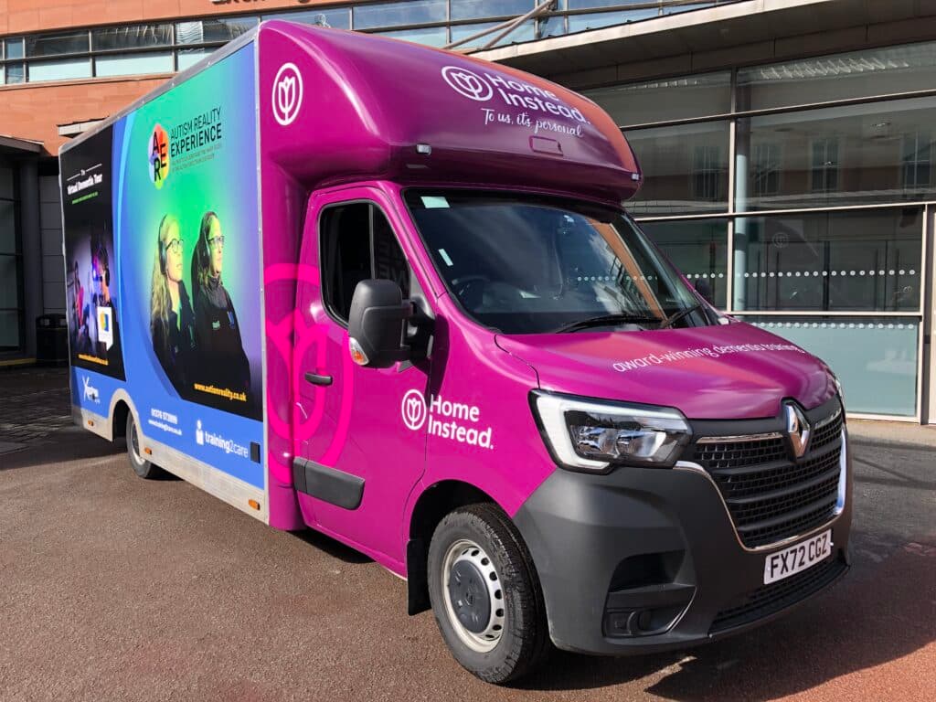 A brightly coloured Home Instead van is parked outside a modern building on a sunny day. - Home Instead