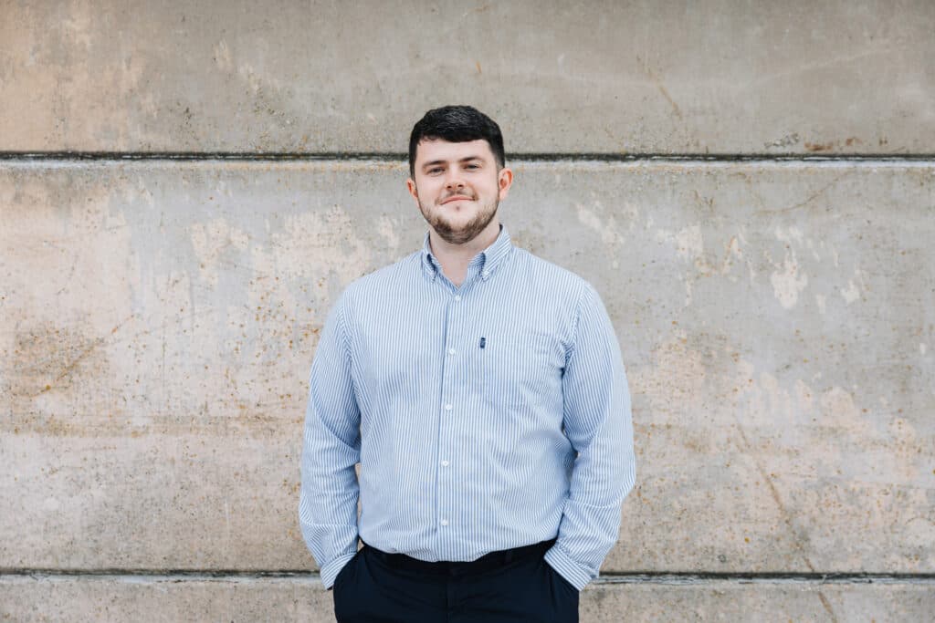 A man in a blue striped shirt stands against a textured concrete wall, hands in his pockets, smiling. - Home Instead