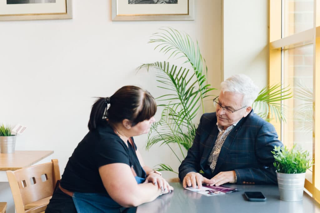 Two people sit at a table in a bright café, having a friendly conversation with documents in front of them. - Home Instead
