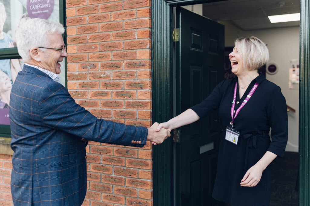 An older man and a woman smile and shake hands at the entrance of a brick building. - Home Instead