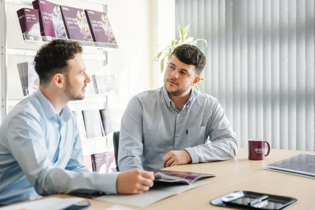 Two men in business attire sit at a desk talking, with documents and a coffee mug in front of them. - Home Instead