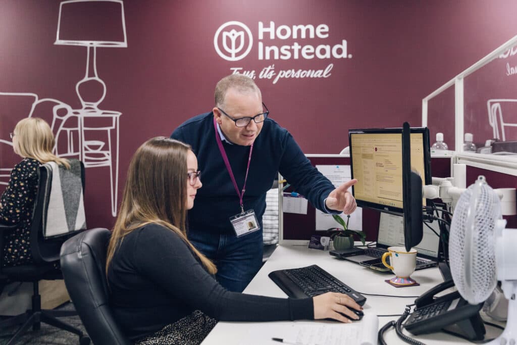 A man stands and points at a computer screen while a woman sits and works in an office. - Home Instead