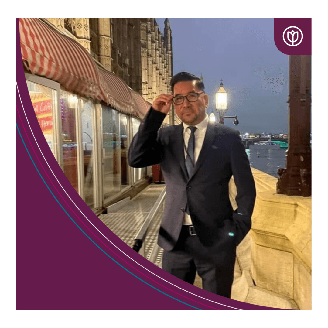 Man in a suit standing by a riverside at dusk, with city lights and historic buildings in the background. - Home Instead
