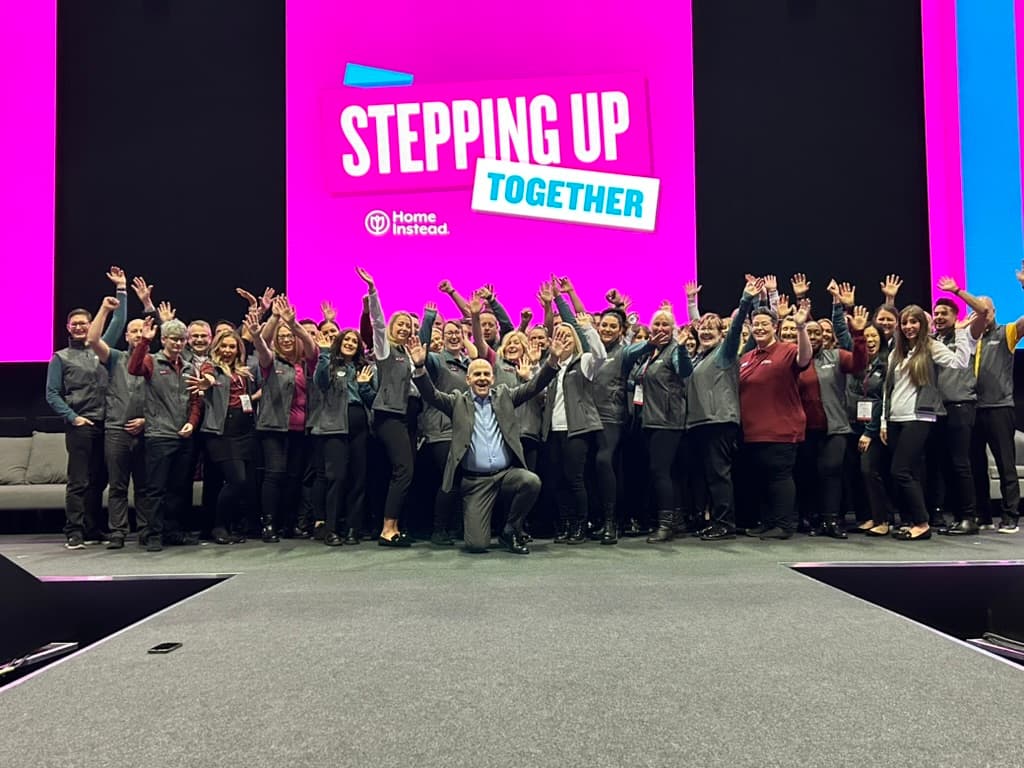 A large group of people pose and wave on stage under a screen that reads "Stepping Up Together. - Home Instead