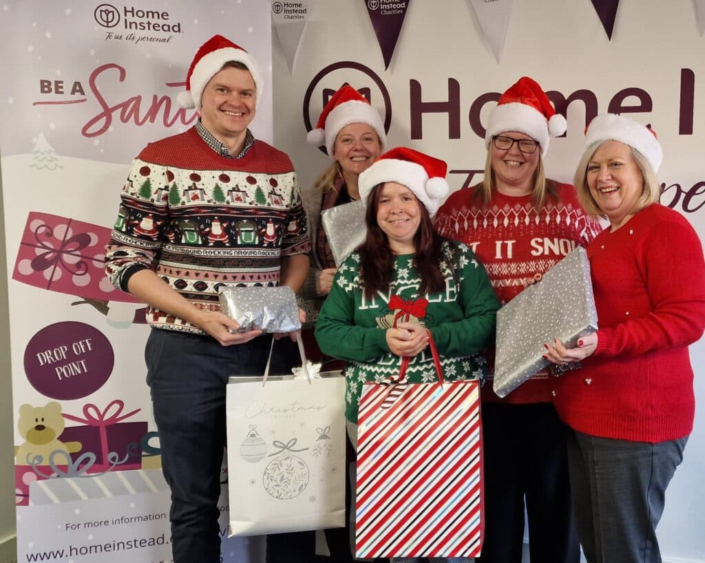 Five people in festive jumpers and Santa hats hold gift bags and presents, smiling in front of a holiday banner. - Home Instead