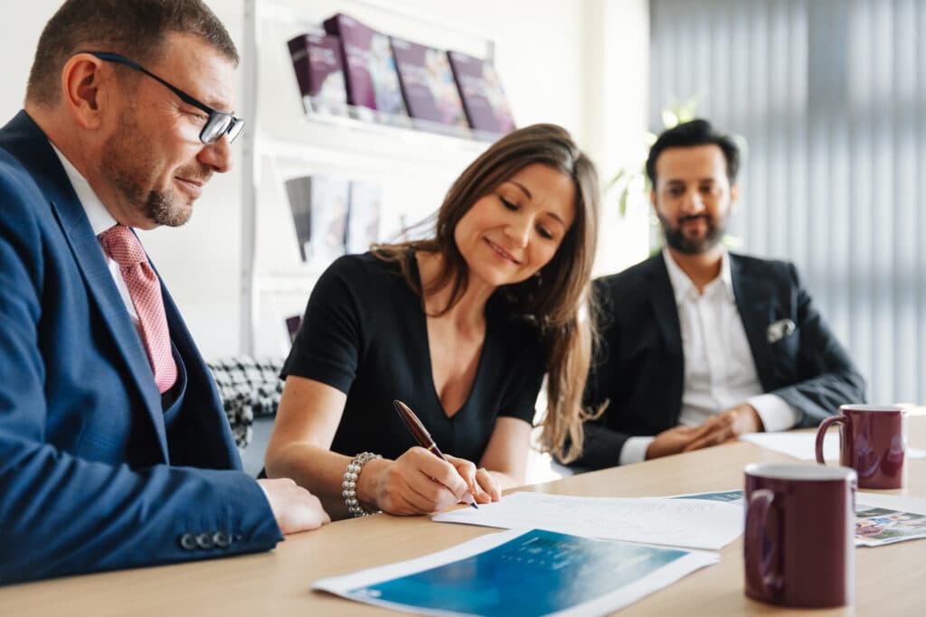 Three people in business attire sit at a table as a woman signs documents, smiling, with two men watching. - Home Instead