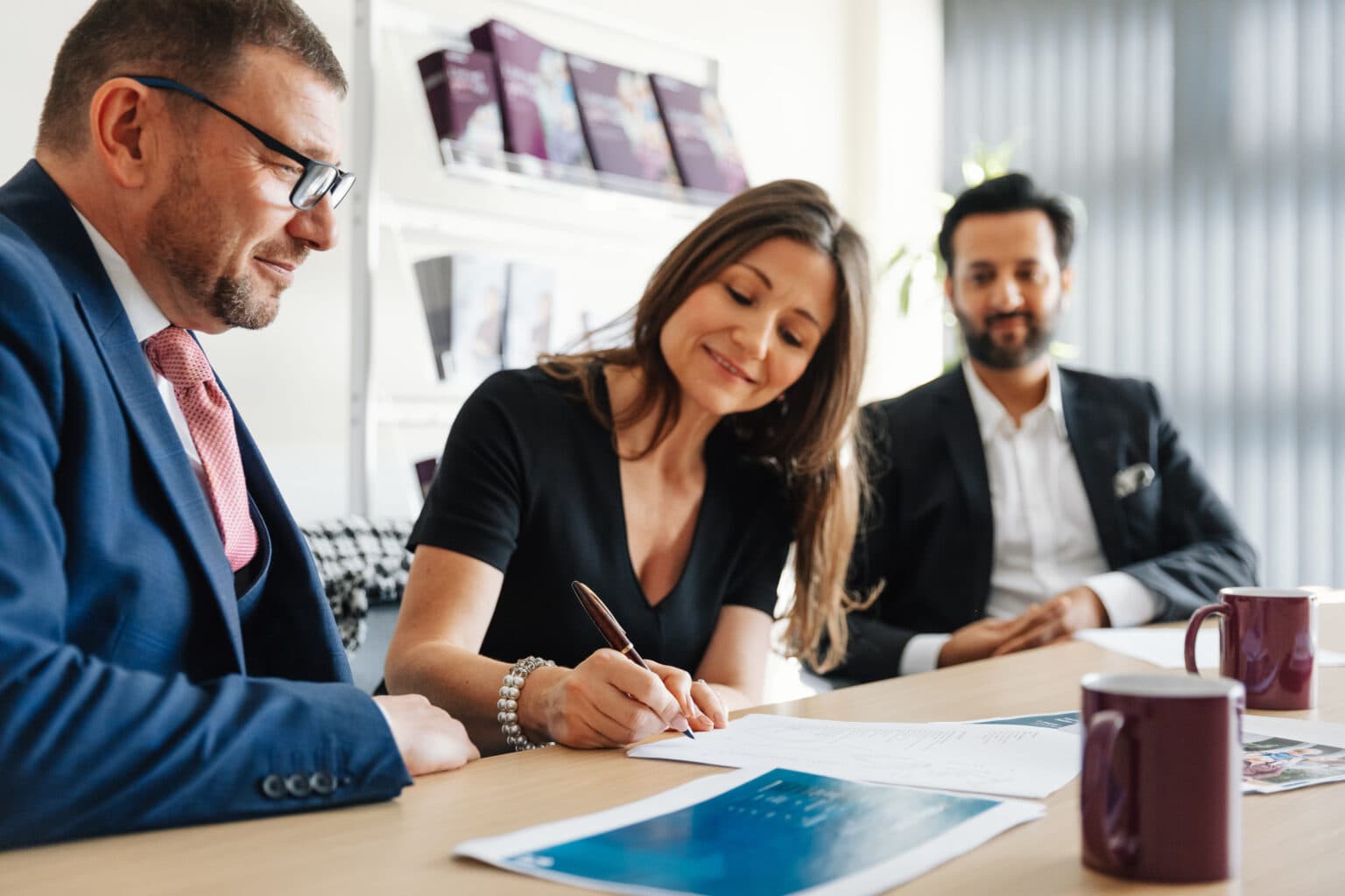 Three people in business attire sit at a table as a woman signs documents, smiling, with two men watching. - Home Instead