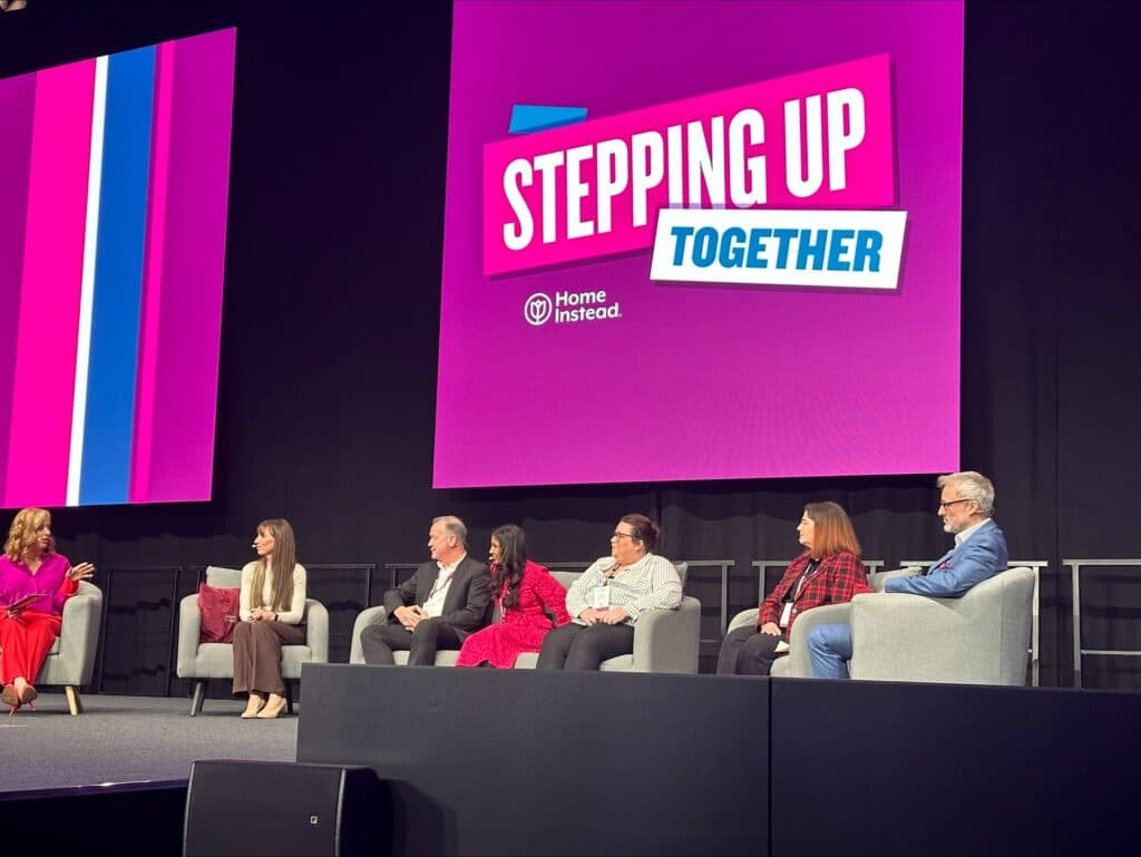 Six people sit on stage with a “Stepping Up Together” sign behind them at a Home Instead event. - Home Instead