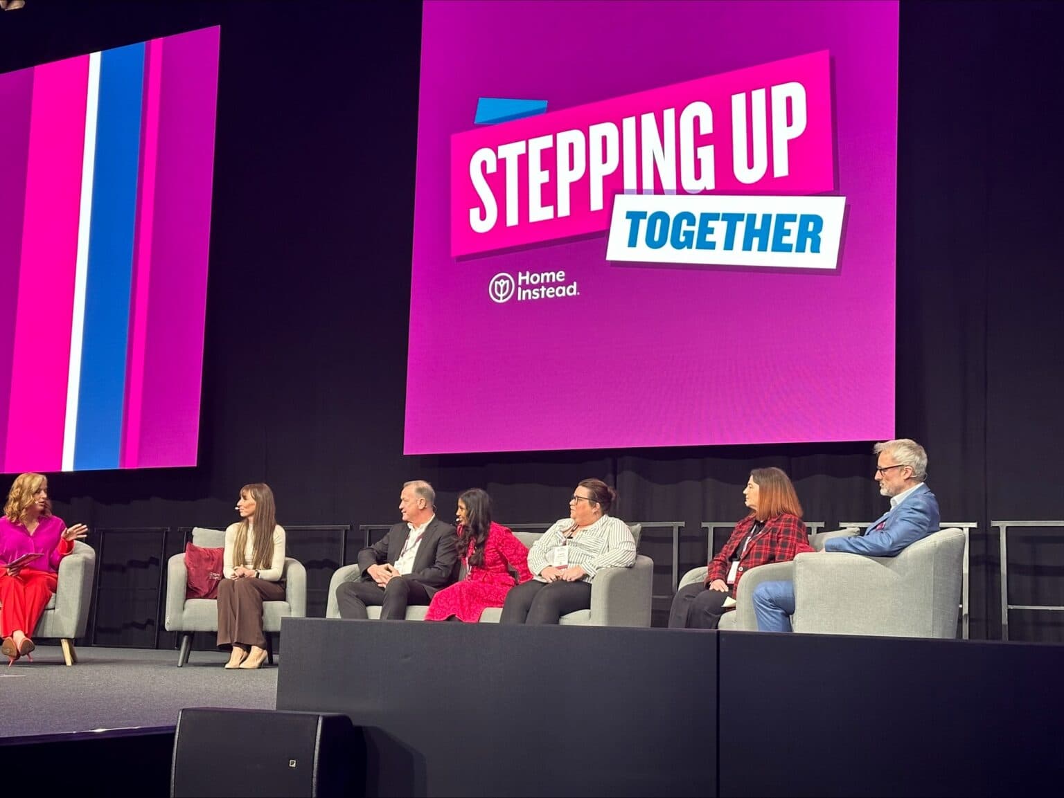 Six people sit on stage with a “Stepping Up Together” sign behind them at a Home Instead event. - Home Instead
