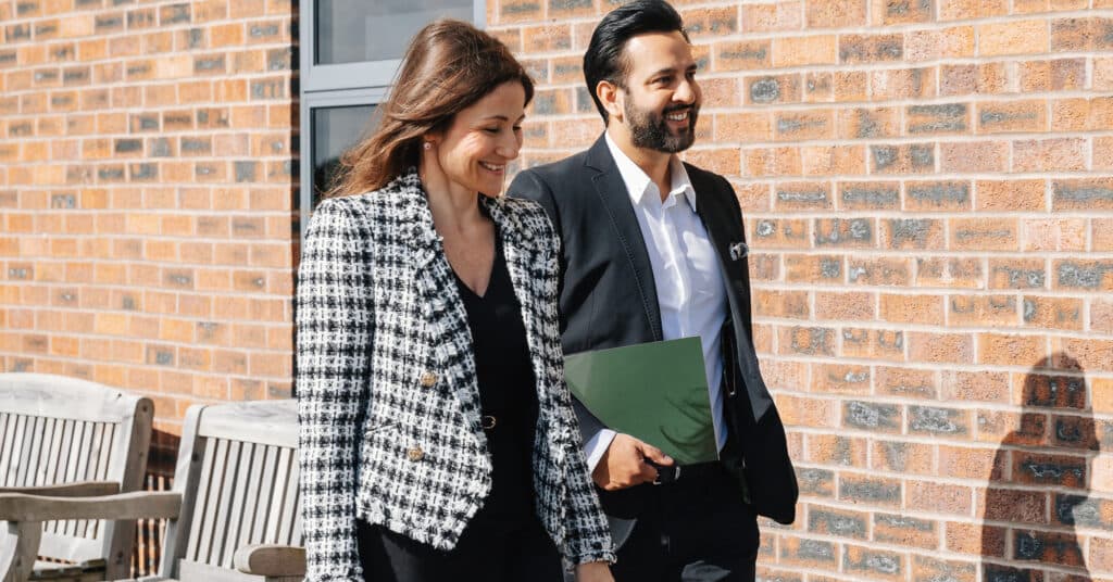 Two professionally dressed people walking outdoors by a brick wall, smiling and talking. - Home Instead