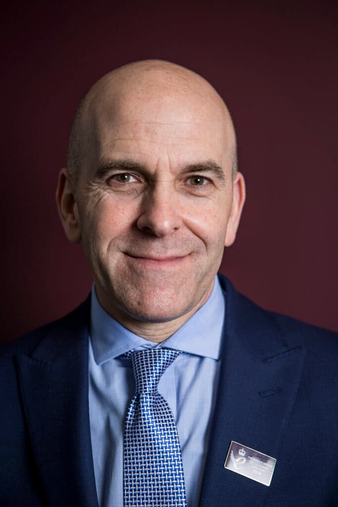 Bald man in a blue suit and tie, smiling, with a name badge, standing in front of a dark red background. - Home Instead