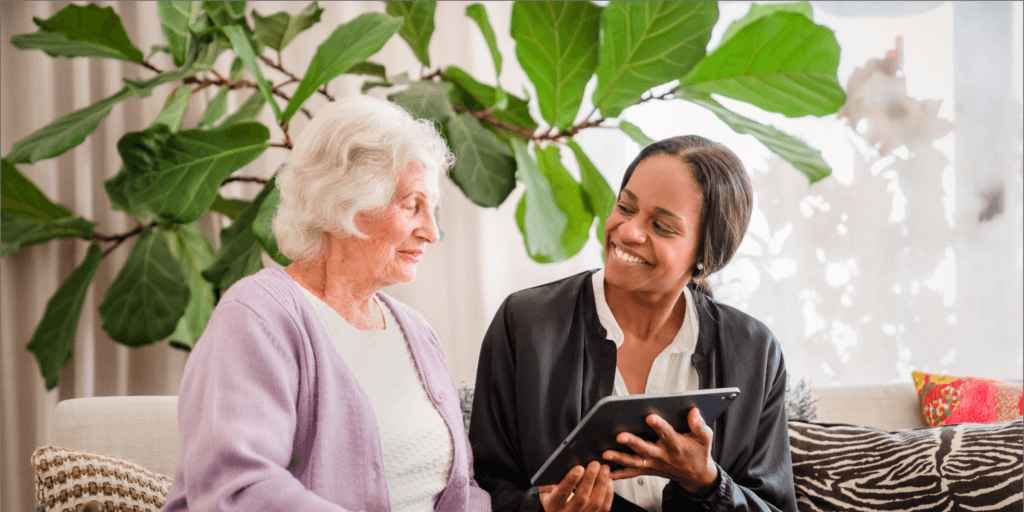 A younger woman shows a tablet to an older woman as they sit together, both smiling warmly. - Home Instead