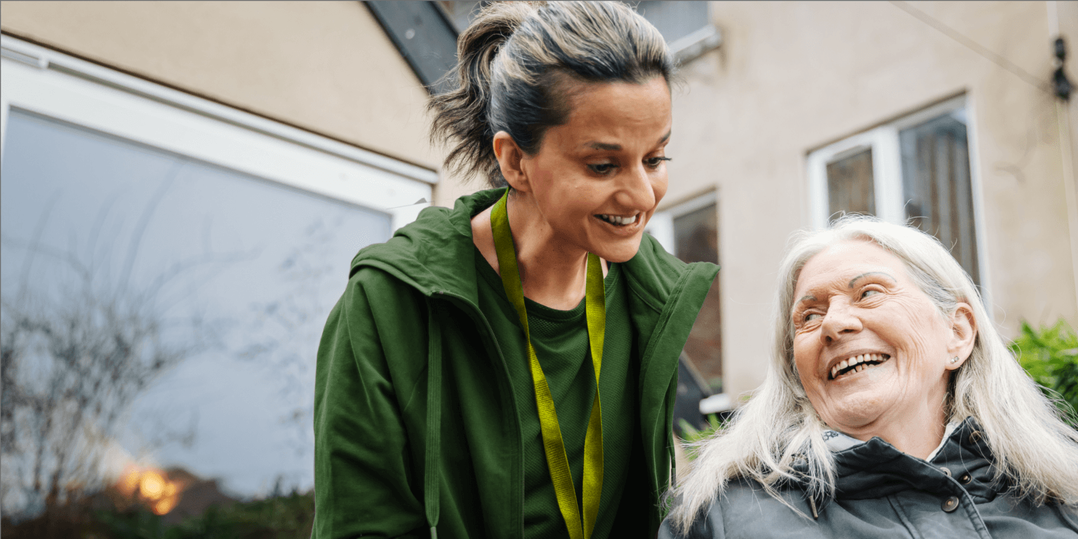 A younger woman shows a tablet to an older woman as they sit together, both smiling warmly. - Home Instead