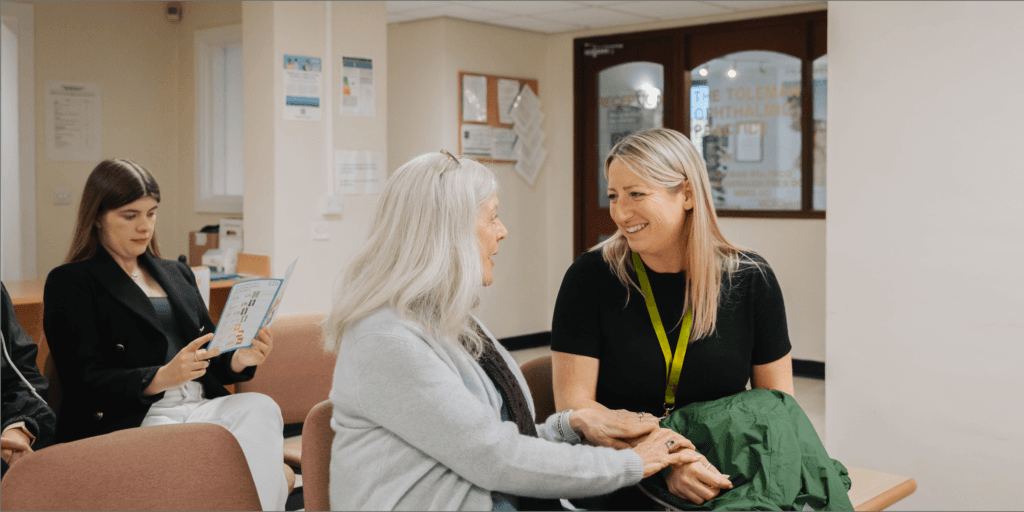 A woman with a lanyard smiles and holds hands with an older woman in a waiting room. - Home Instead