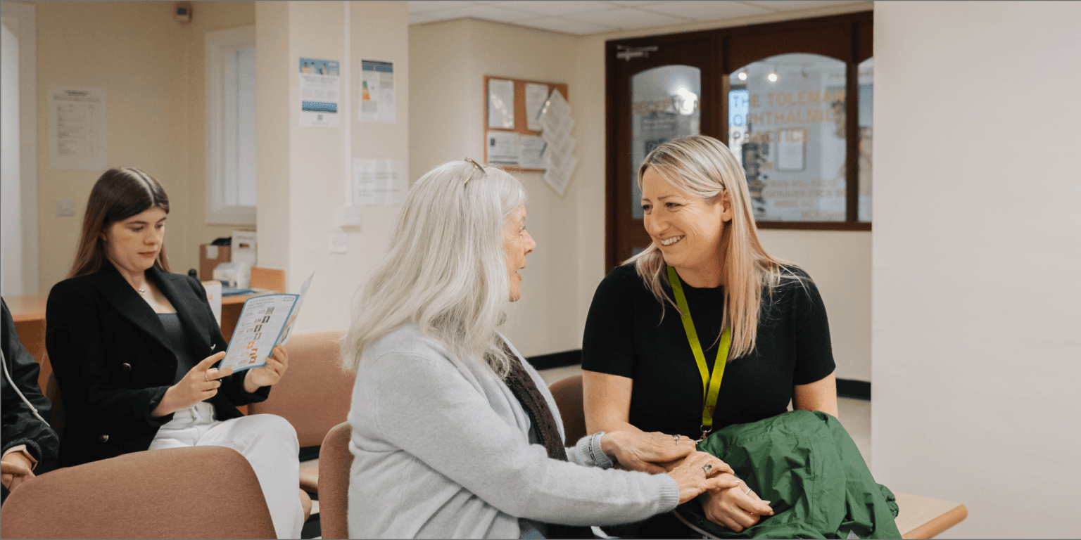 A woman with a lanyard smiles and holds hands with an older woman in a waiting room. - Home Instead