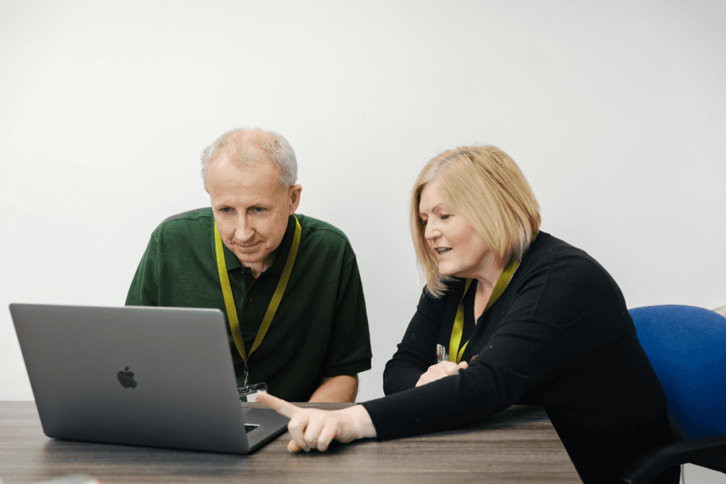 Two colleagues with lanyards look at a laptop together; the woman points at the screen. - Home Instead