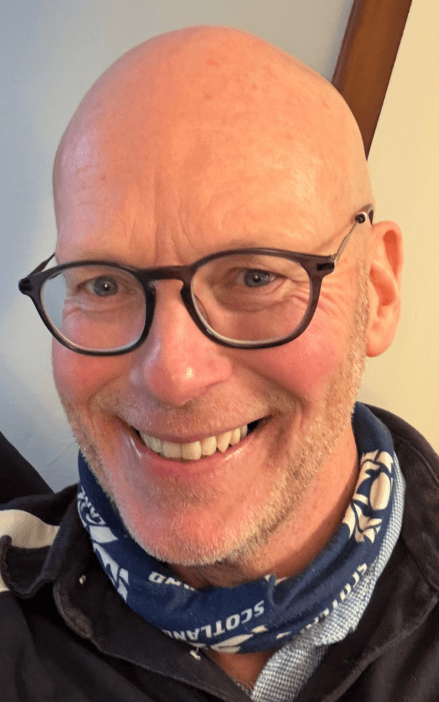 A smiling bald man with glasses and a blue scarf with white patterns. - Home Instead