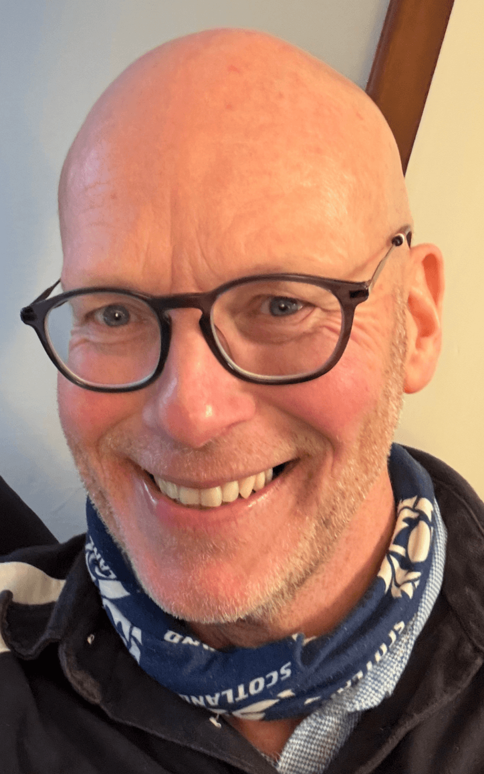 A smiling bald man with glasses and a blue scarf with white patterns. - Home Instead