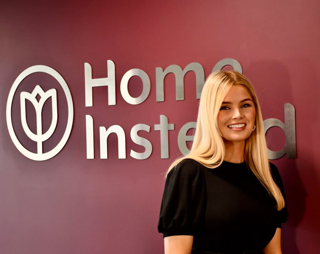 Smiling woman stands in front of a maroon wall with a "Home Instead" logo and text. - Home Instead
