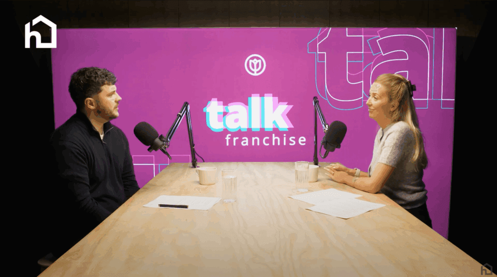 Two people sit at a podcast table with microphones, talking in front of a purple "talk franchise" backdrop. - Home Instead