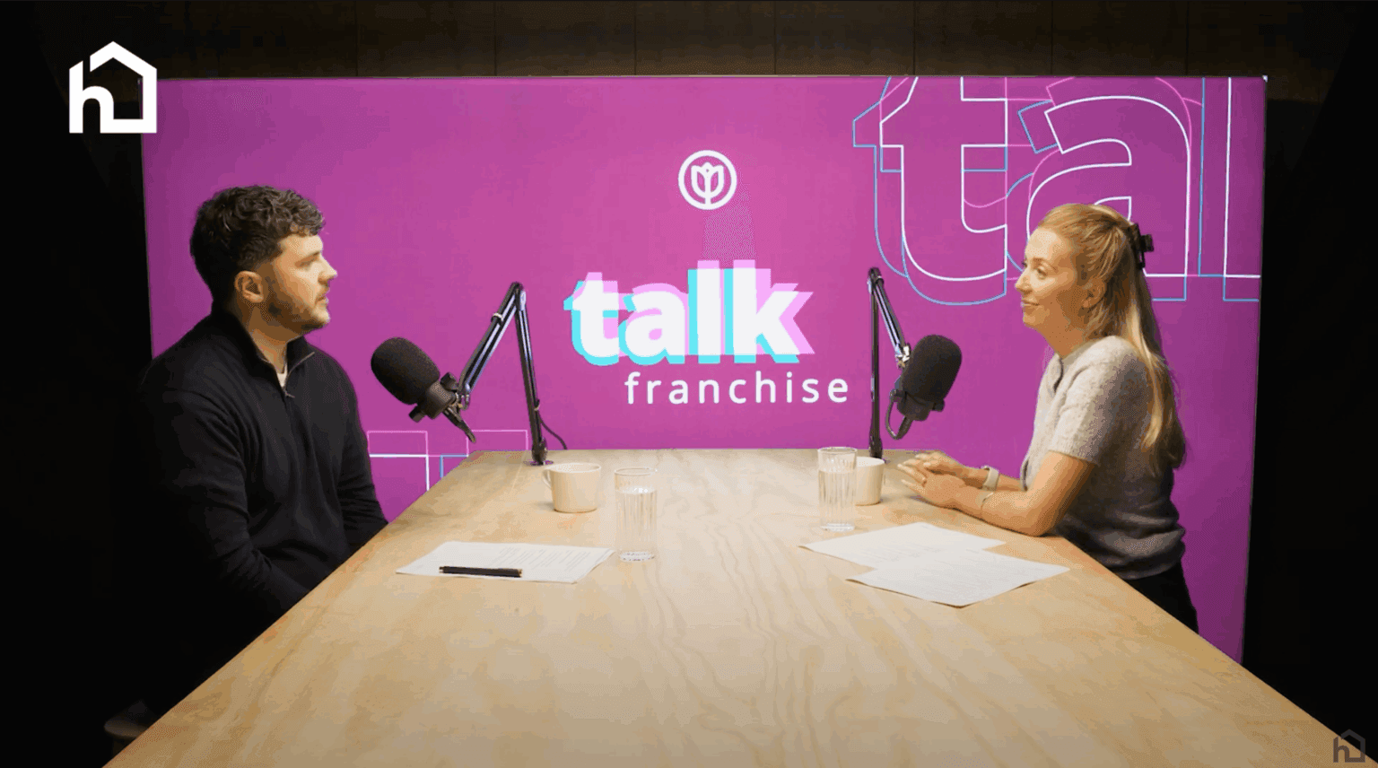 Two people sit at a podcast table with microphones, talking in front of a purple "talk franchise" backdrop. - Home Instead
