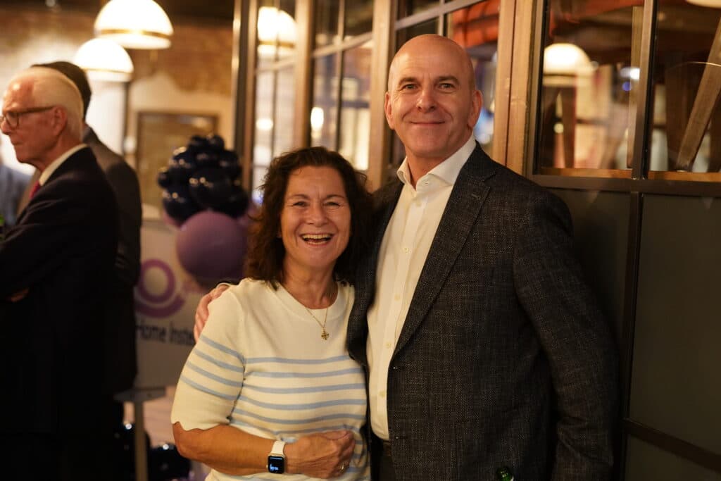 A smiling woman and man pose together at an indoor social event with people in the background. - Home Instead