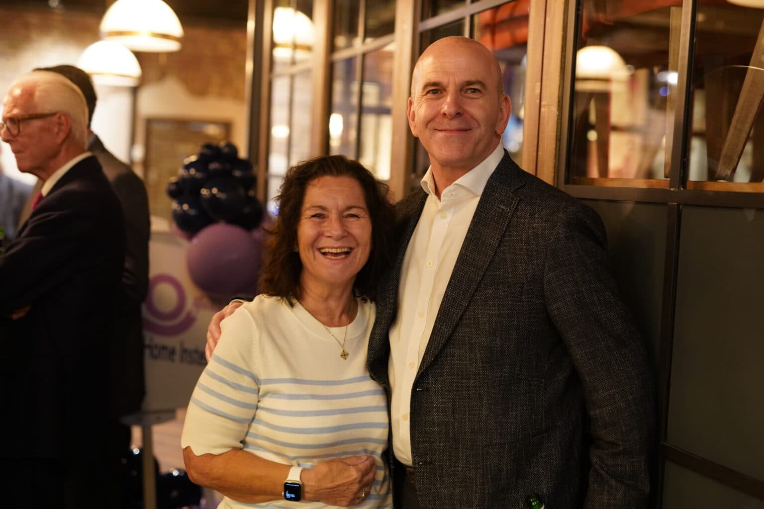 A smiling woman and man pose together at an indoor social event with people in the background. - Home Instead