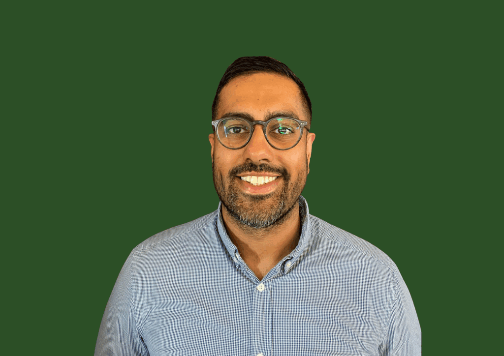 A man wearing glasses and a blue shirt smiles in front of a solid green background. - Home Instead