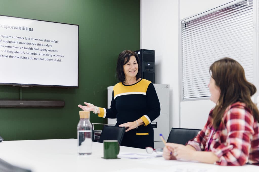 Woman giving a presentation to a seated woman in an office meeting room with a screen displaying text. - Home Instead