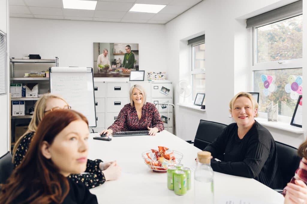 Five women sit around a meeting table in a brightly lit office, with snacks and drinks on the table. - Home Instead