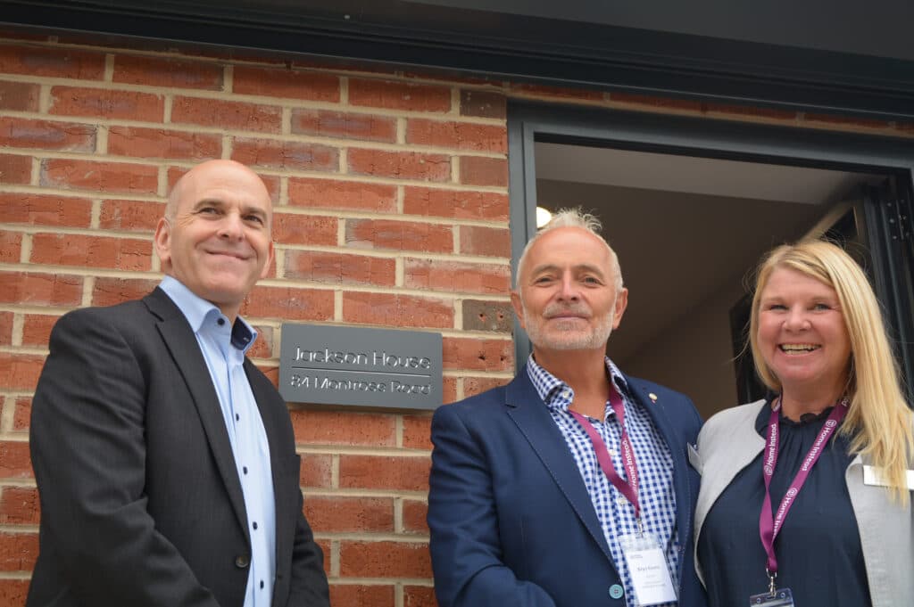 Three people smiling and standing outside a brick building next to a sign that reads "Jackson House". - Home Instead