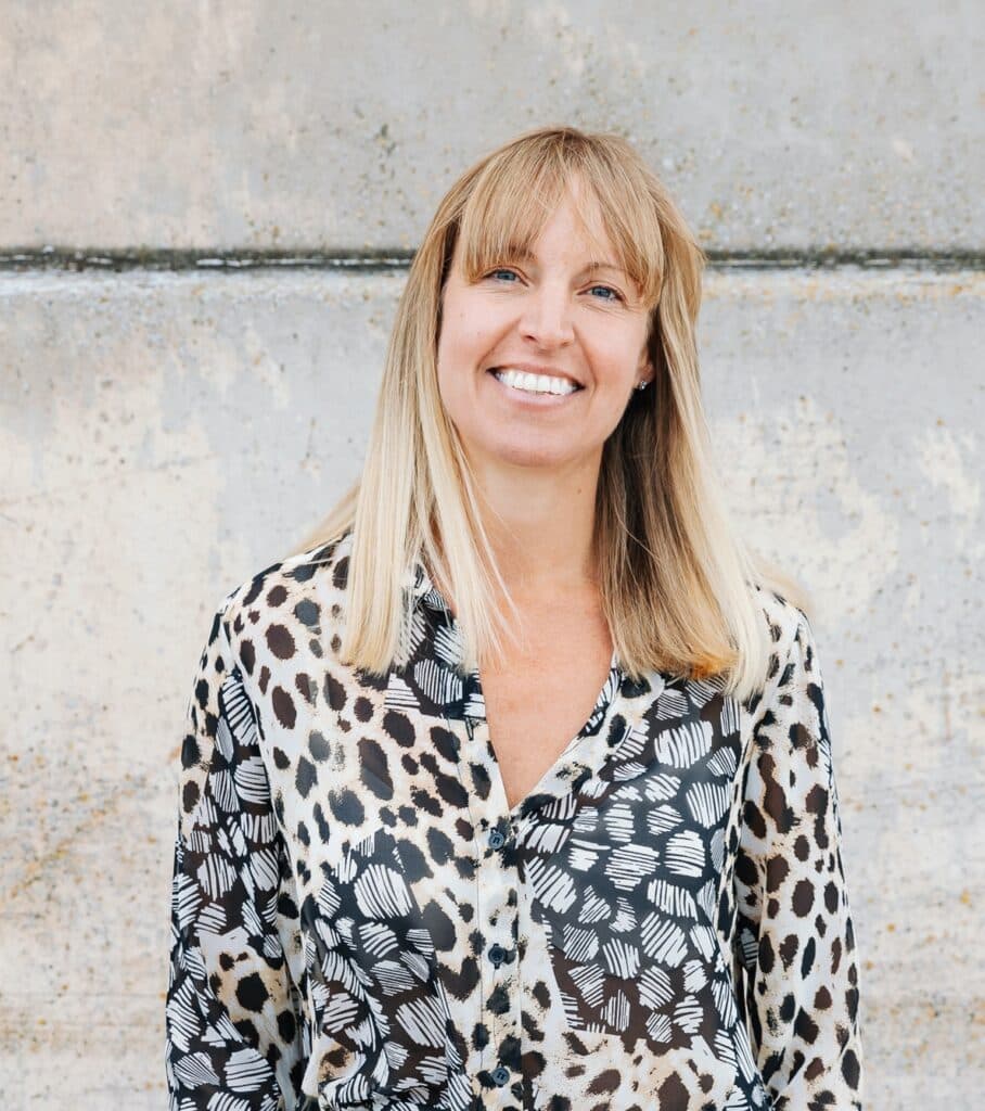 Woman with blonde hair in a patterned shirt smiles in front of a textured concrete wall. - Home Instead