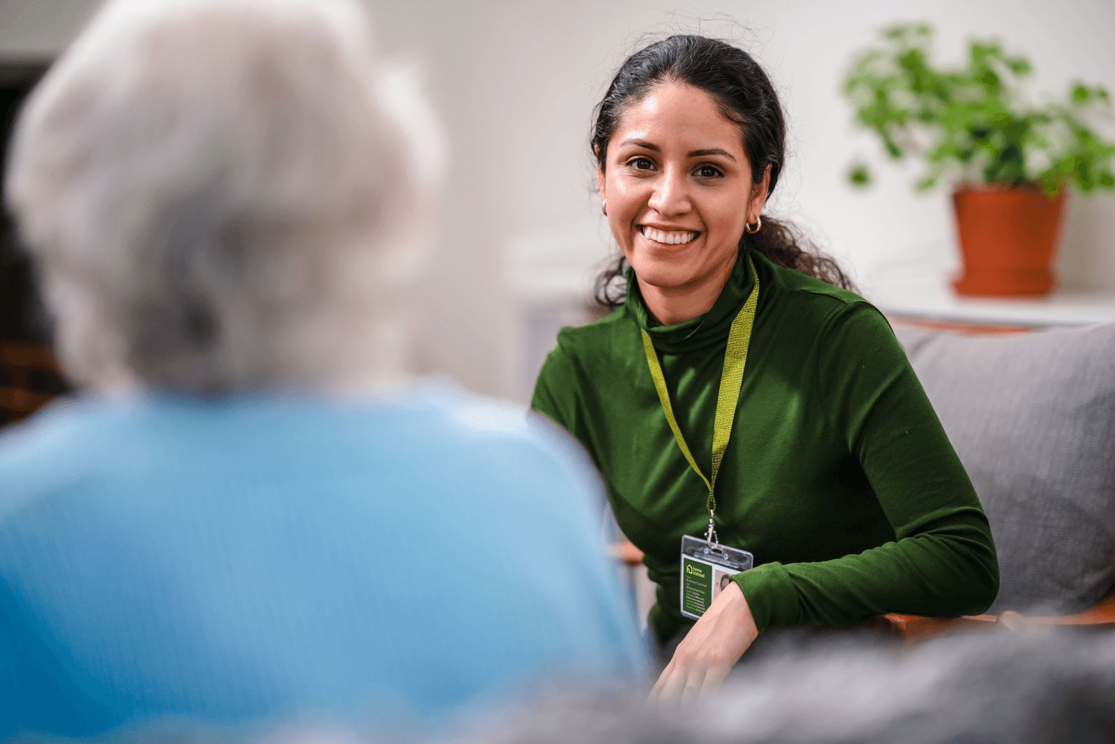 A smiling woman with a badge talks to an older person seated opposite her in a cosy room. - Home Instead