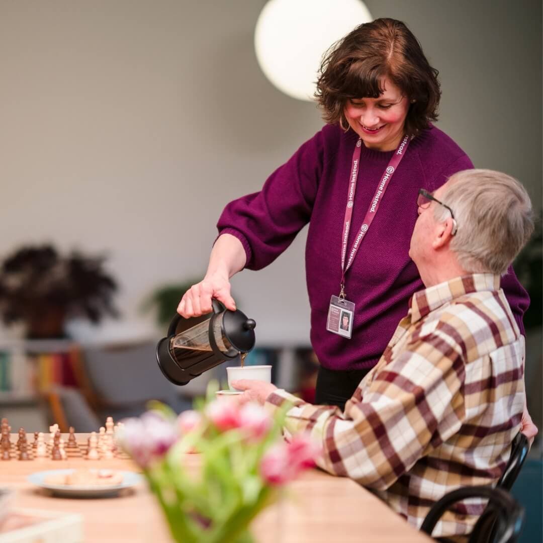 A woman pours coffee for an elderly man sitting at a table, with a chessboard and flowers in the foreground. - Home Instead