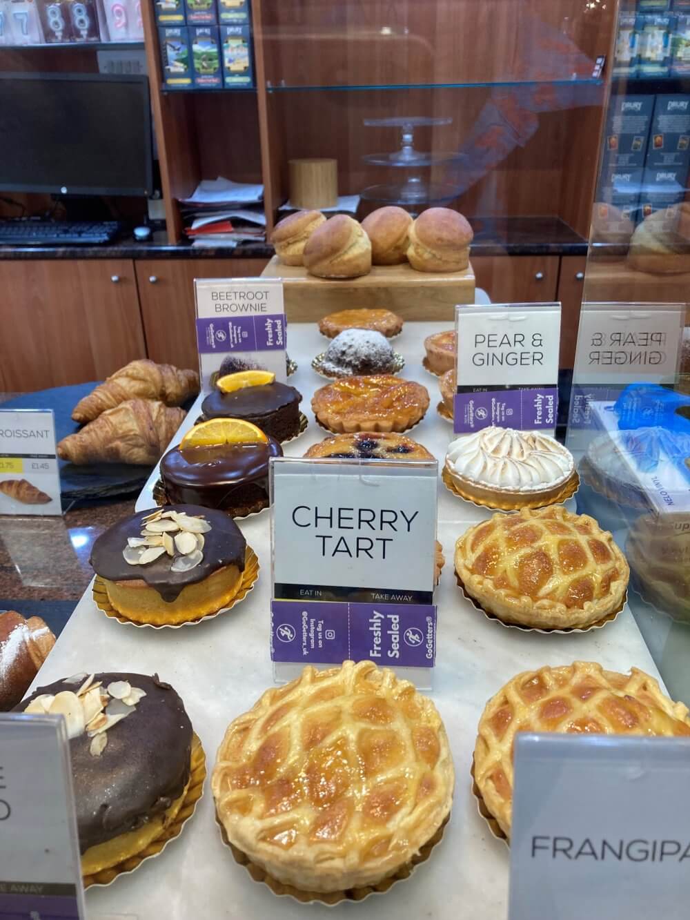 A bakery display showing various pastries, including cherry tart, pear and ginger tart, and beetroot brownie. - Home Instead