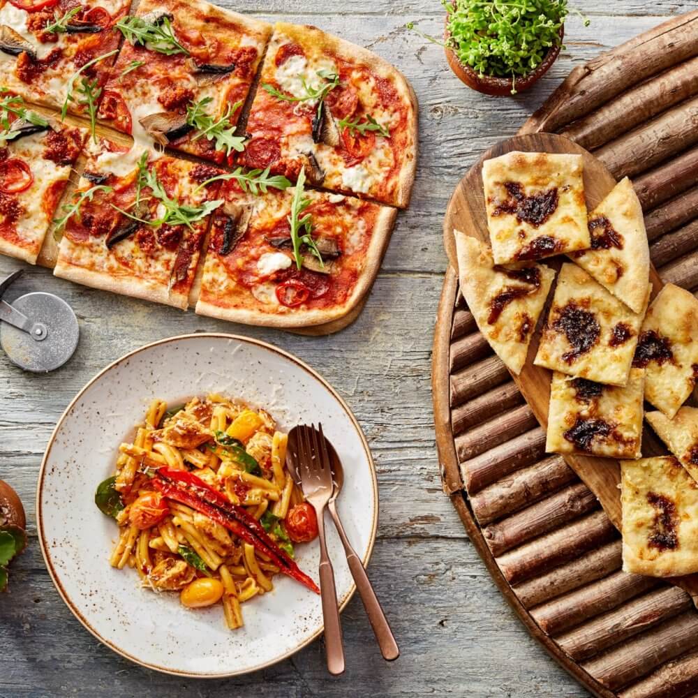 A table with pizza, pasta, and breadsticks garnished with herbs and vegetables on wooden boards. - Home Instead