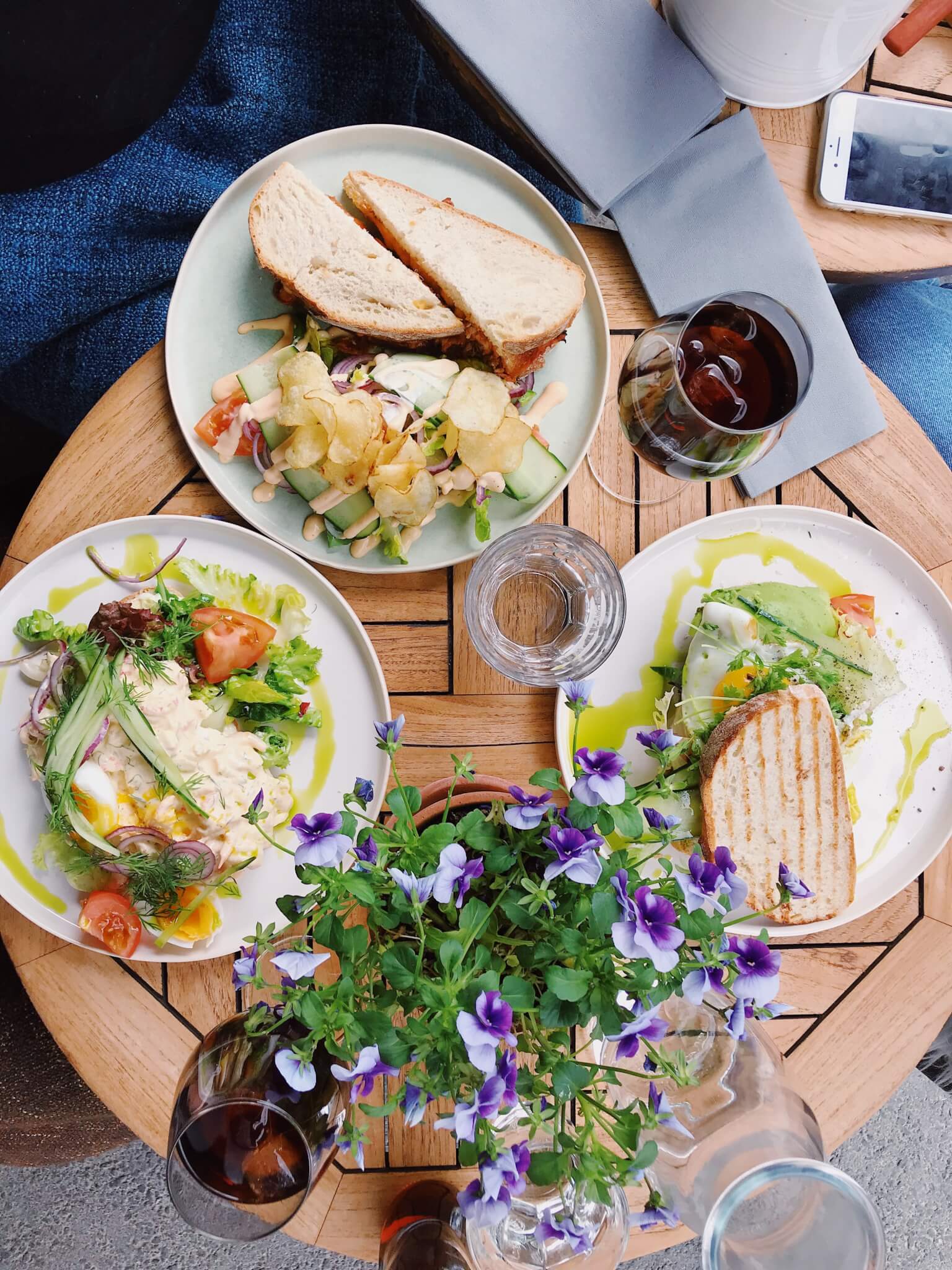 A table with three plates of food including sandwiches and salads, two glasses of red wine, and a vase of purple flowers. - Home Instead