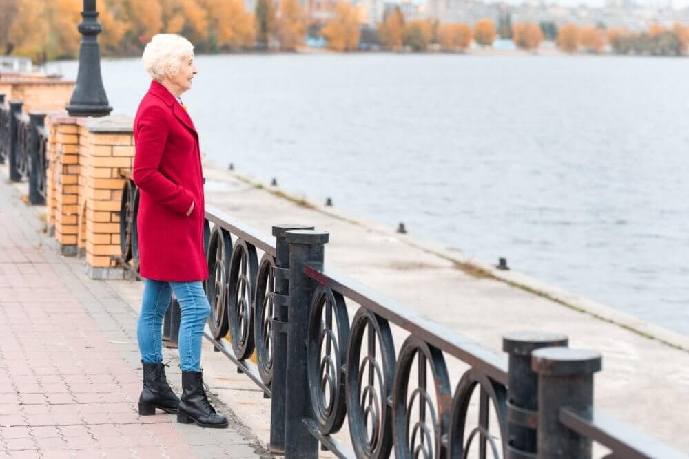 Elderly woman in red coat and blue jeans stands by a waterfront railing, gazing at the river on an autumn day. - Home Instead Bournemouth & Christchurch