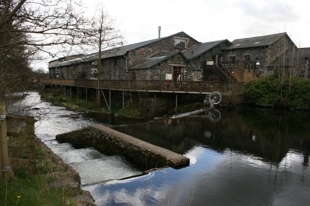 A stone building with a wooden deck over a calm river with trees and a small bridge in the foreground. - Home Instead