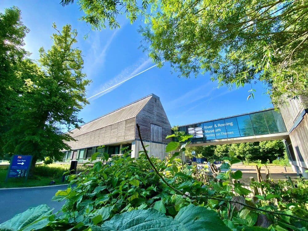 Modern building surrounded by greenery on a sunny day, with a sign reading "River & Rowing Museum" on a glass bridge. - Home Instead