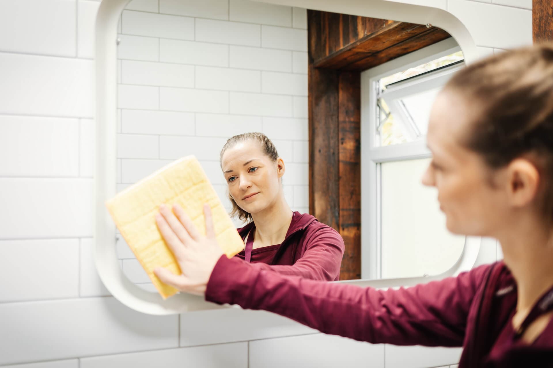A woman in a maroon top cleans a bathroom mirror with a yellow cloth. - Home Instead
