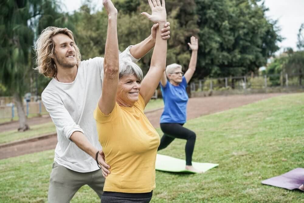 Man assisting elderly woman with yoga pose in a park, another elderly woman practicing in the background. - Home Instead