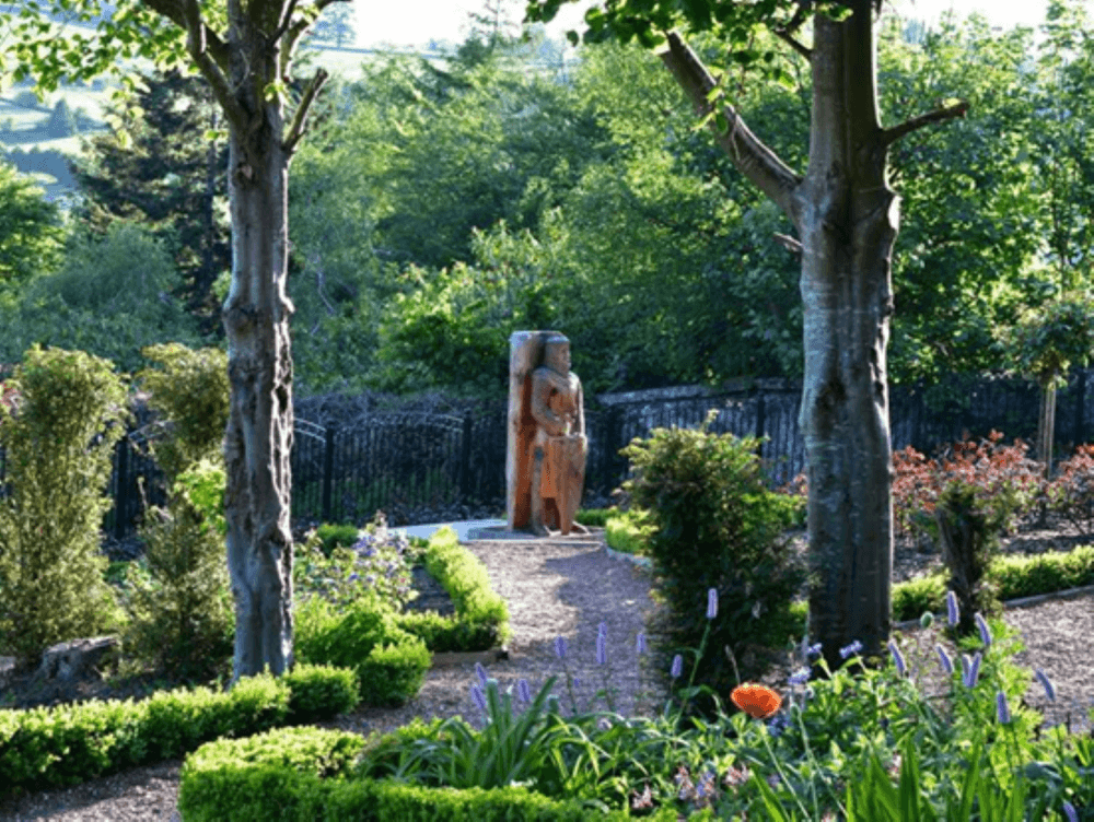 A lush garden with various plants, pathways, and a tall wooden sculpture surrounded by trees. - Home Instead