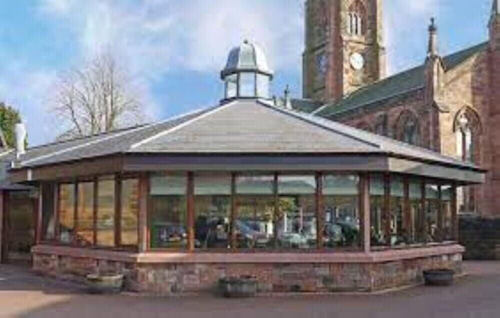 A modern glass pavilion with a gray roof near an old stone church with a clock tower on a clear day. - Home Instead