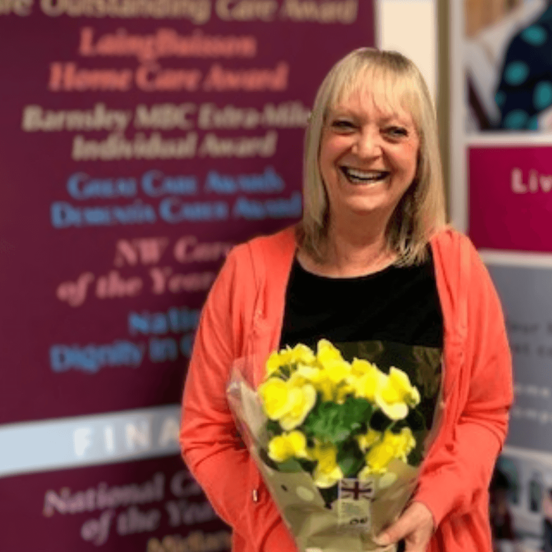 A smiling woman holds a bouquet of yellow flowers, standing in front of a backdrop with various award titles. - Home Instead