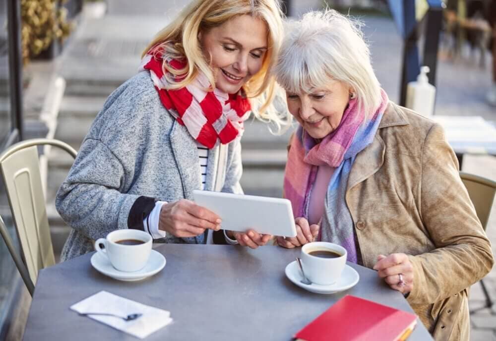 Two women sitting at a table outside, smiling and looking at a tablet, with two cups of coffee in front of them. - Home Instead