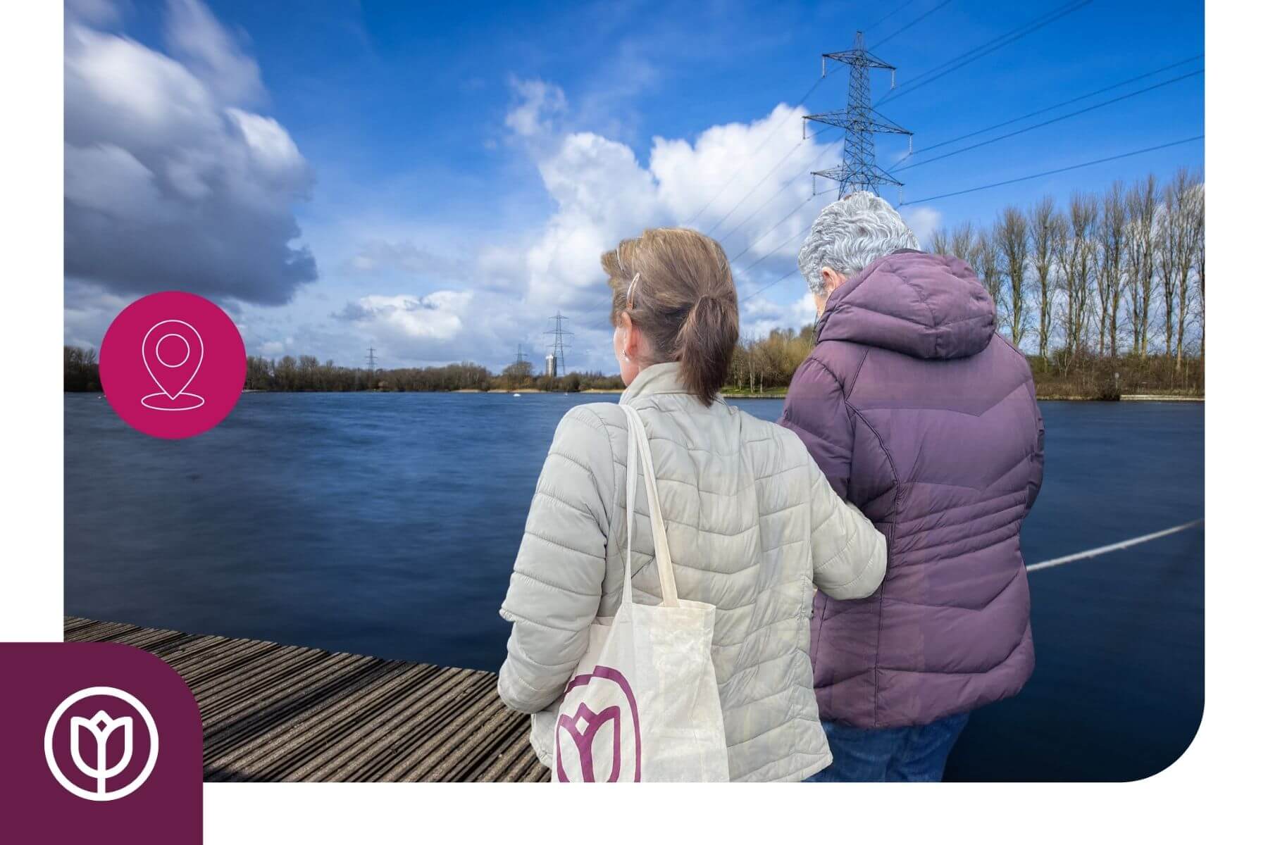 Two people in jackets standing by a lake, with one holding a tote bag. Power lines and trees are in the background. - Home Instead