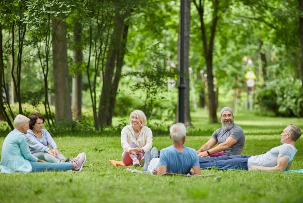 A group of older adults sit on yoga mats in a park, engaging in a relaxed conversation surrounded by greenery. - Home Instead Southampton