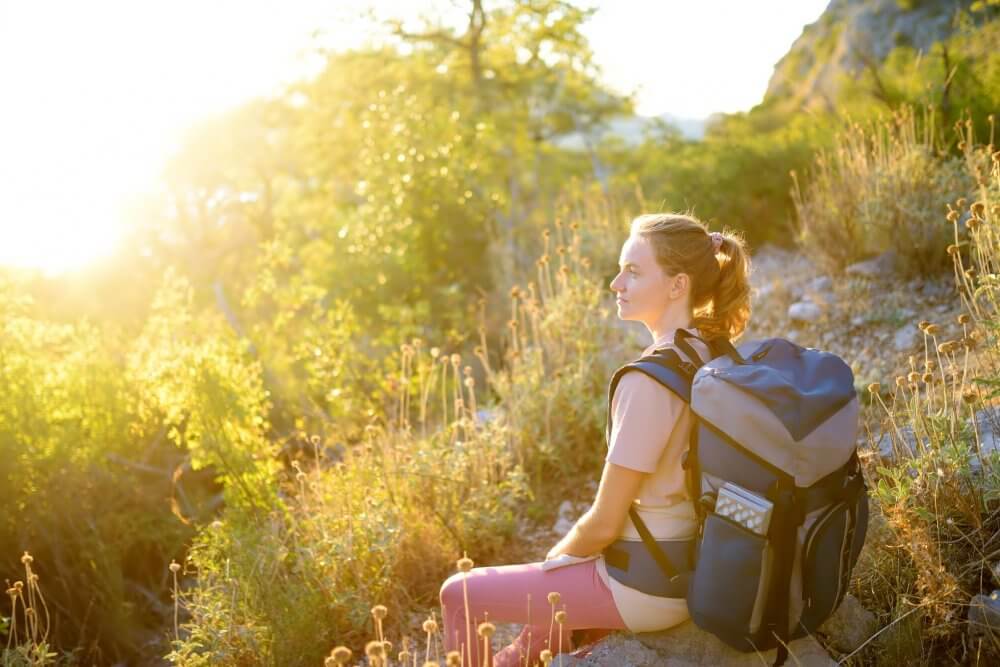 Woman in hiking gear sits on a rock, basking in the sun, surrounded by greenery and dry plants. - Home Instead
