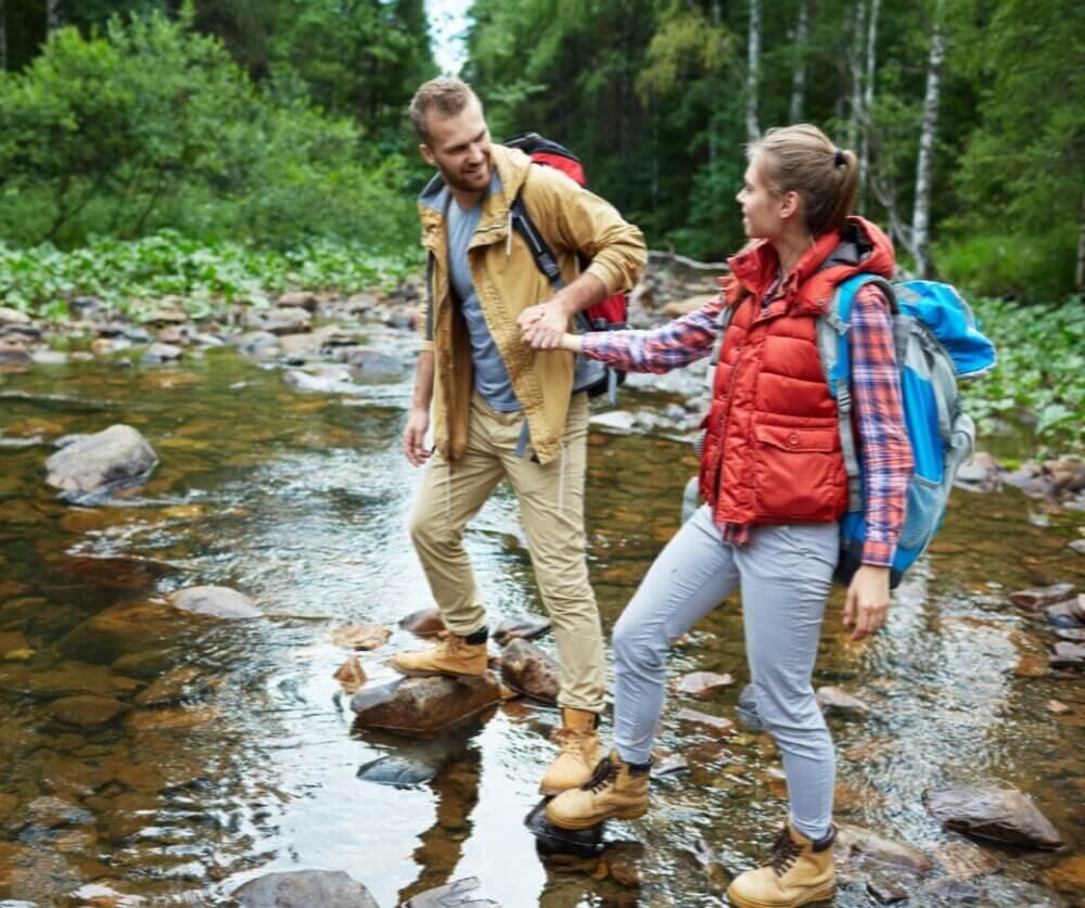 Two hikers in outdoor gear help each other cross a shallow stream surrounded by lush greenery. - Home Instead